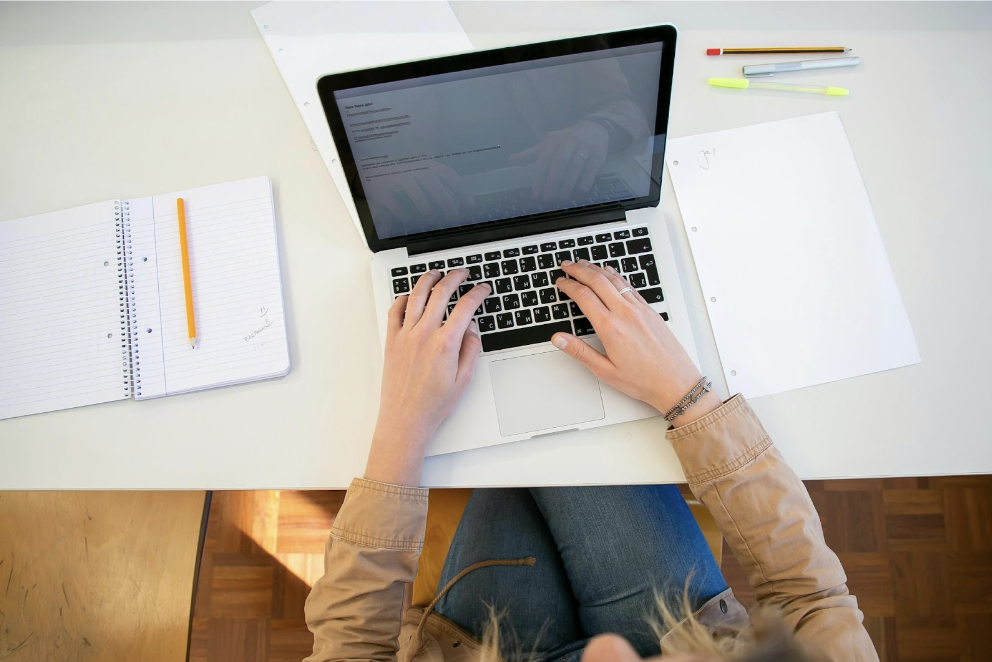 A person works on a laptop with a notebook and paper and writing utensils on a white desktop.