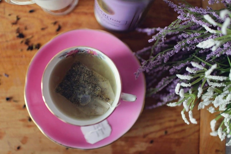 A teacup with a teabag full of natural components sits next to a cluster of flowers. 