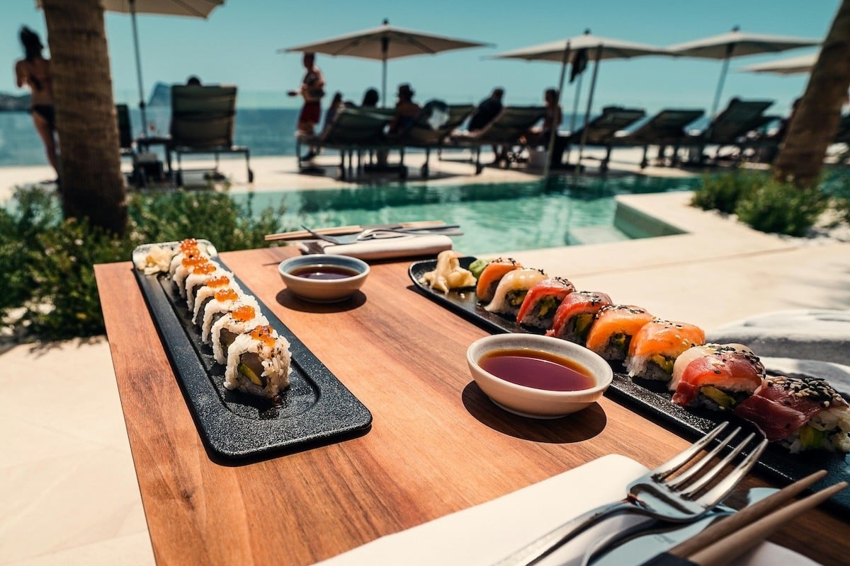 A tray of sushi with sauces sits on a table in front of a pool and beach.jpg