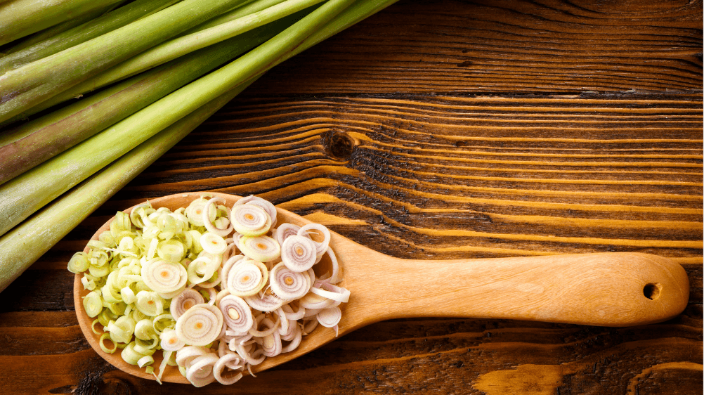 A wooden spoon holding sliced lemongrass ends next to several full stalks of lemongrass