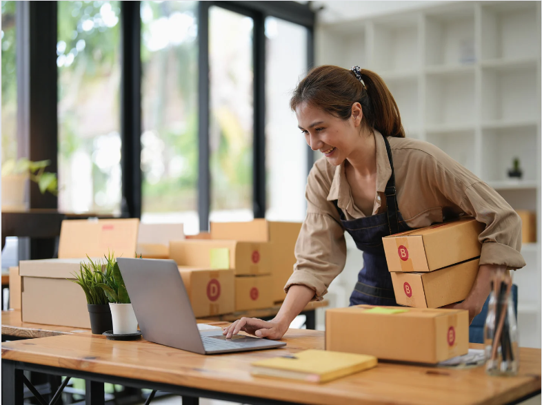 a woman works on a laptop while holding boxes in a small storefront