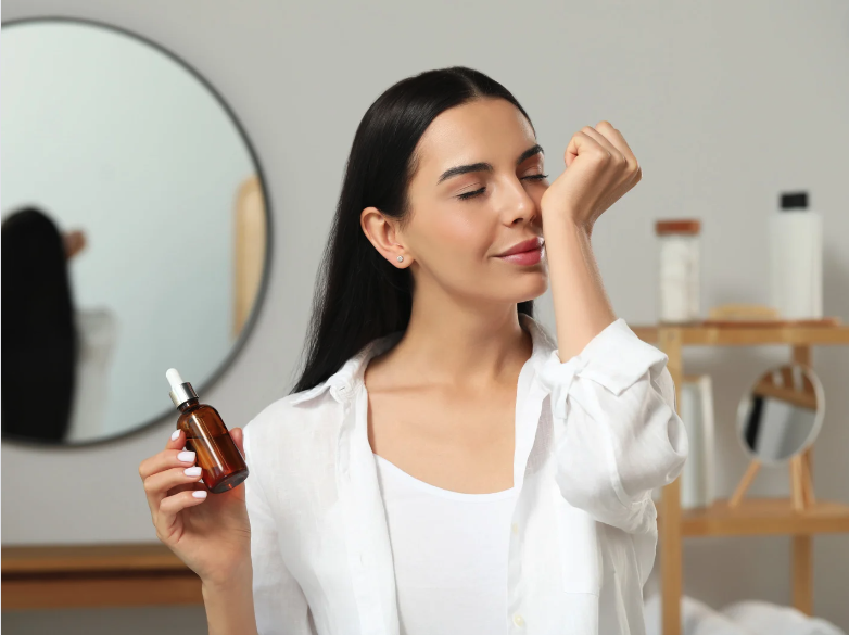 a woman in her bathroom holds a bottle of essential oil and sniffs the scent from her wrist